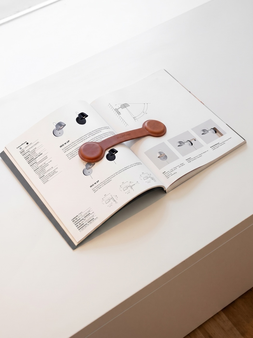 An open Martinez catalog spread for the IRIO wall light, a leather sample tag resting on top.